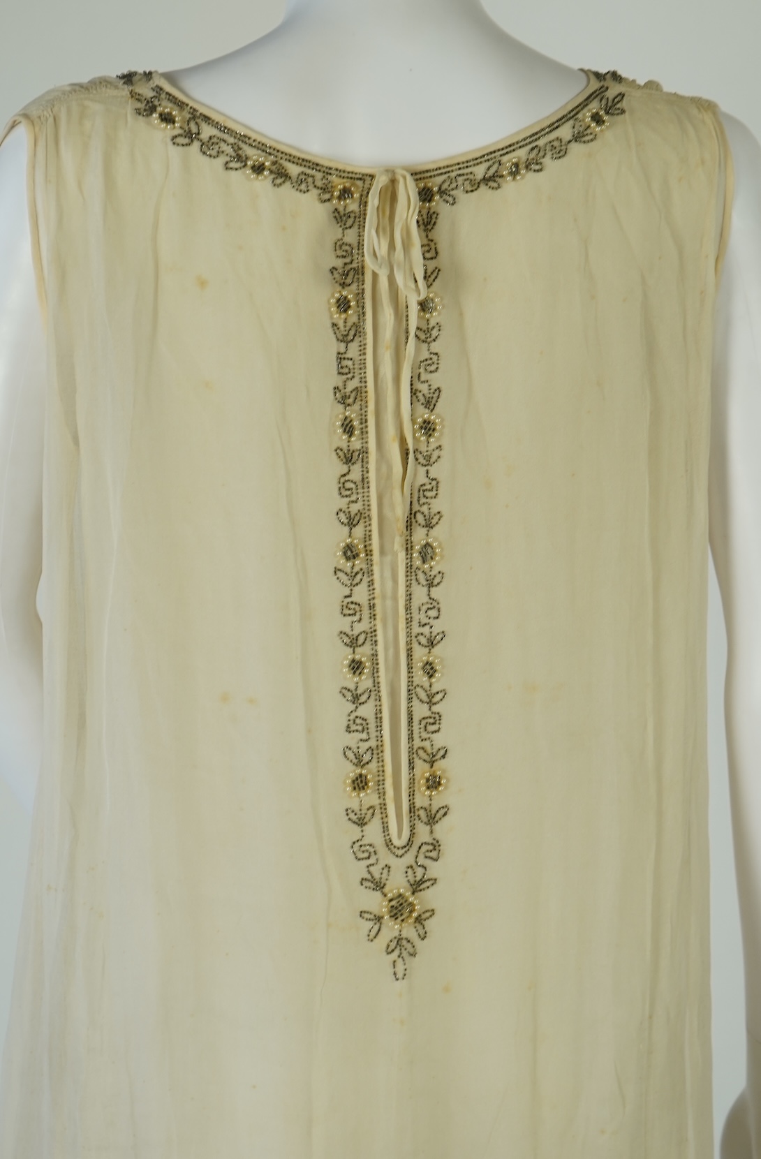 A 1920's cream silk georgette three quarter length, silk lined ‘flapper dress’. Designed with beaded and pearl floral embroidery, a stylish long beaded keyhole slit at the back, a beaded front bodice and flying beaded pa